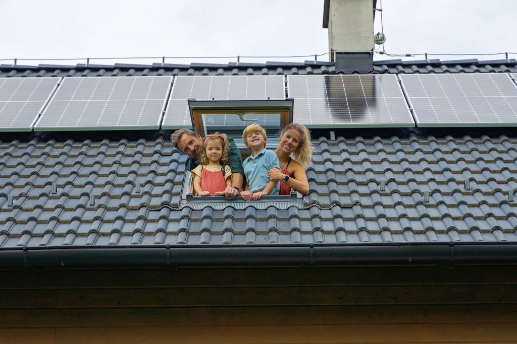 Happy family leaning out from skylight window in their house with solar panels on the roof.